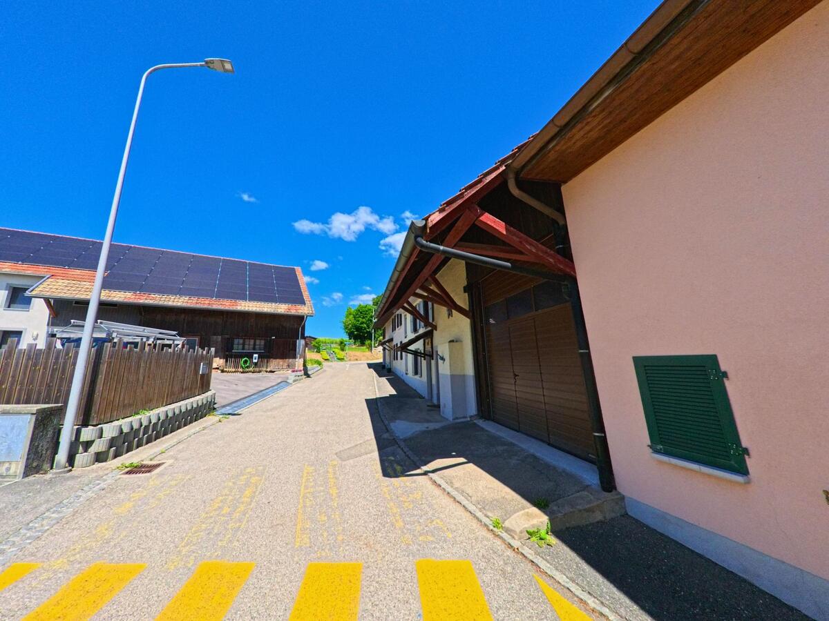 In Alle (2942, Kanton Jura) fuehrt eine schmale Seitenstrasse an Wirtschaftsgebaeuden vorbei, darunter eine Scheune mit Solarpanelen auf dem Dach.
