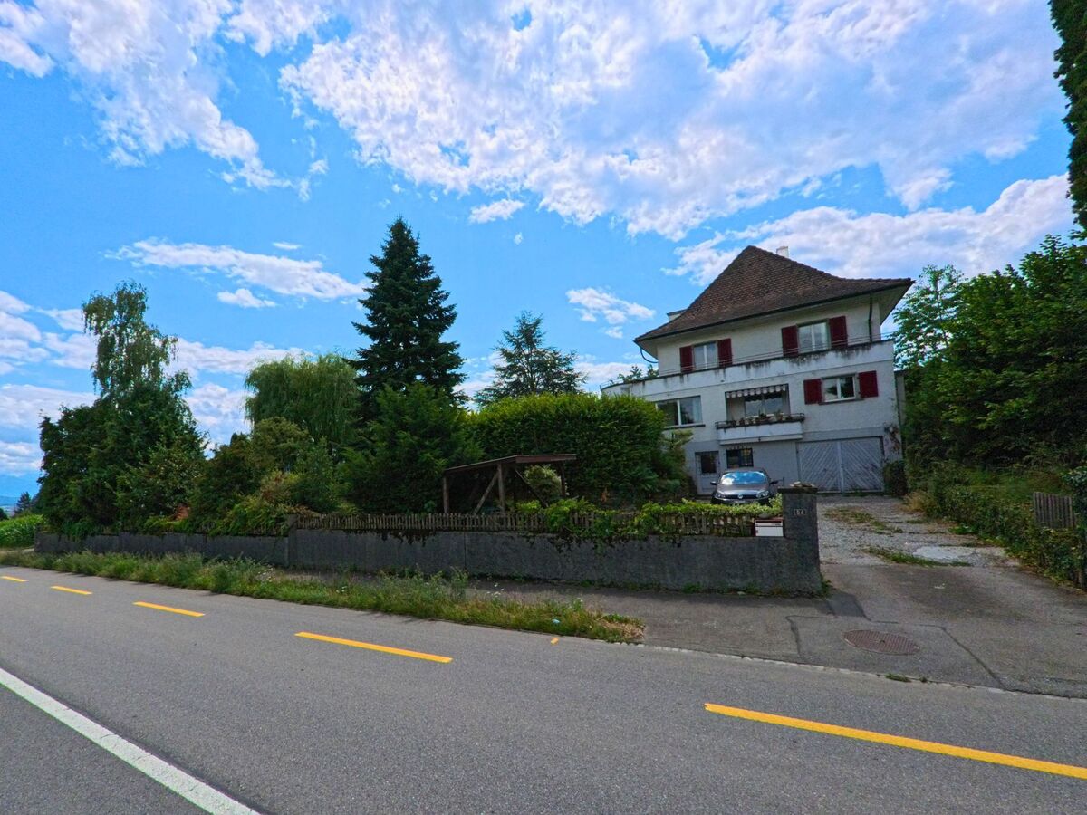 In Allmendingen (3112, Kanton Bern) liegt ein freistehendes Wohnhaus mit roten Fensterläden und grossem Garten am Rand der Landstrasse, eingebettet in dichte Vegetation.