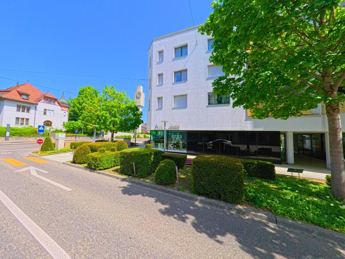 In Allschwil (4123, Kanton Baselland) trifft eine Nebenstrasse mit Zebrastreifen auf einen Platz mit modernen Gebäuden, gepflegten Sträuchern und einem Uhrenturm im Hintergrund.