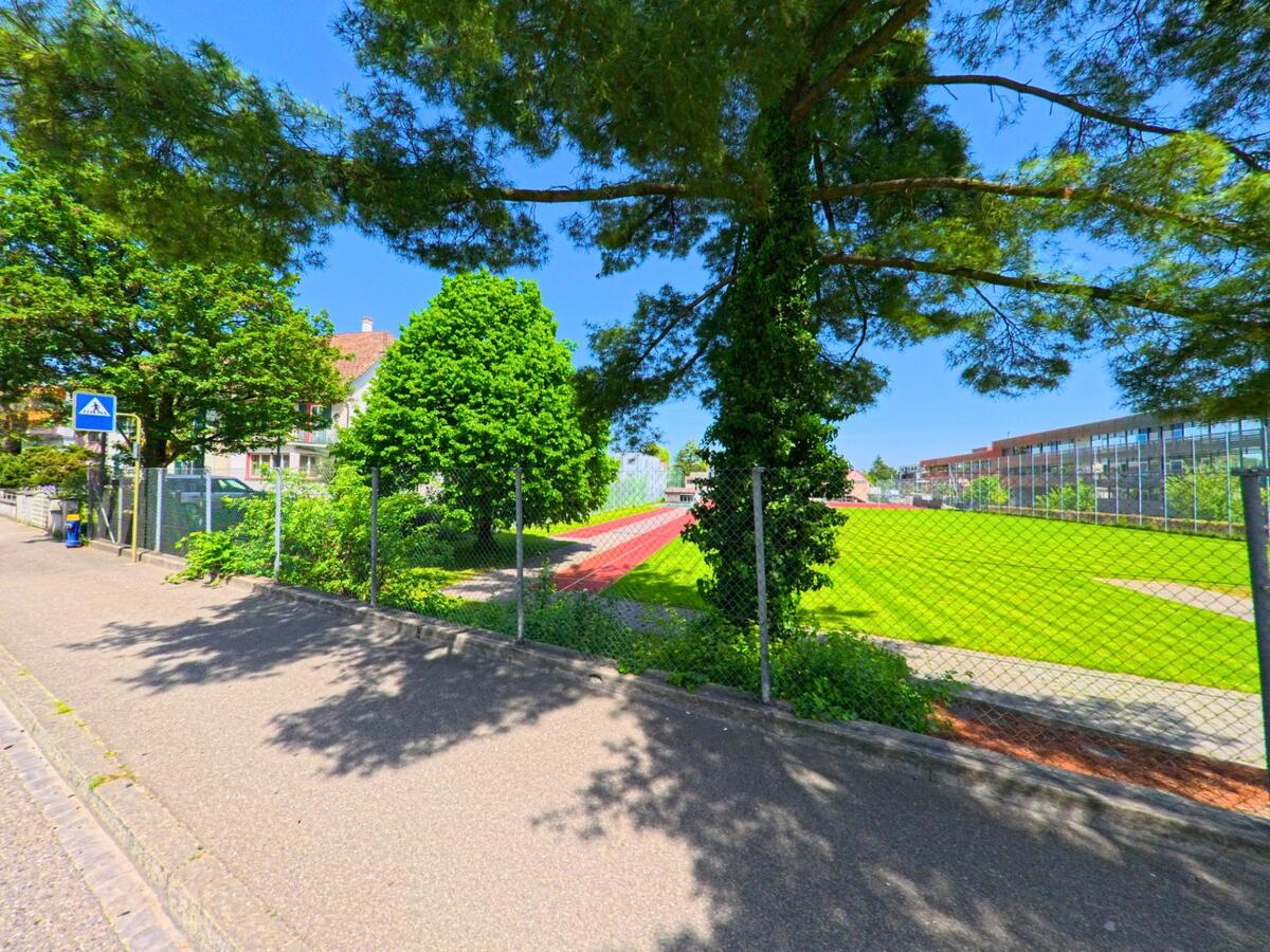 In Allschwil (4123, Kanton Baselland) verläuft ein Gehweg entlang eines umzäunten Sportplatzes mit Laufbahn und angrenzender moderner Schulanlage.