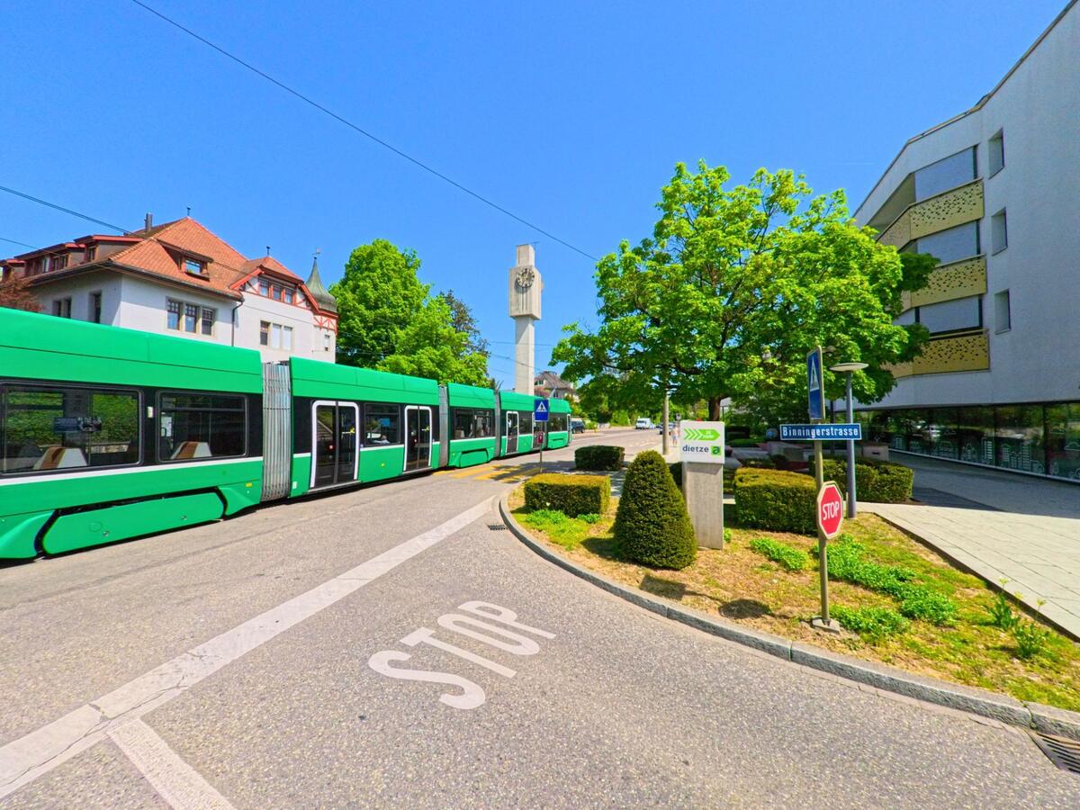 In Allschwil (4123, Kanton Baselland) überquert eine grüne Tramlinie die Binningerstrasse in einem urbanen Bereich mit modernen und historischen Gebäuden.