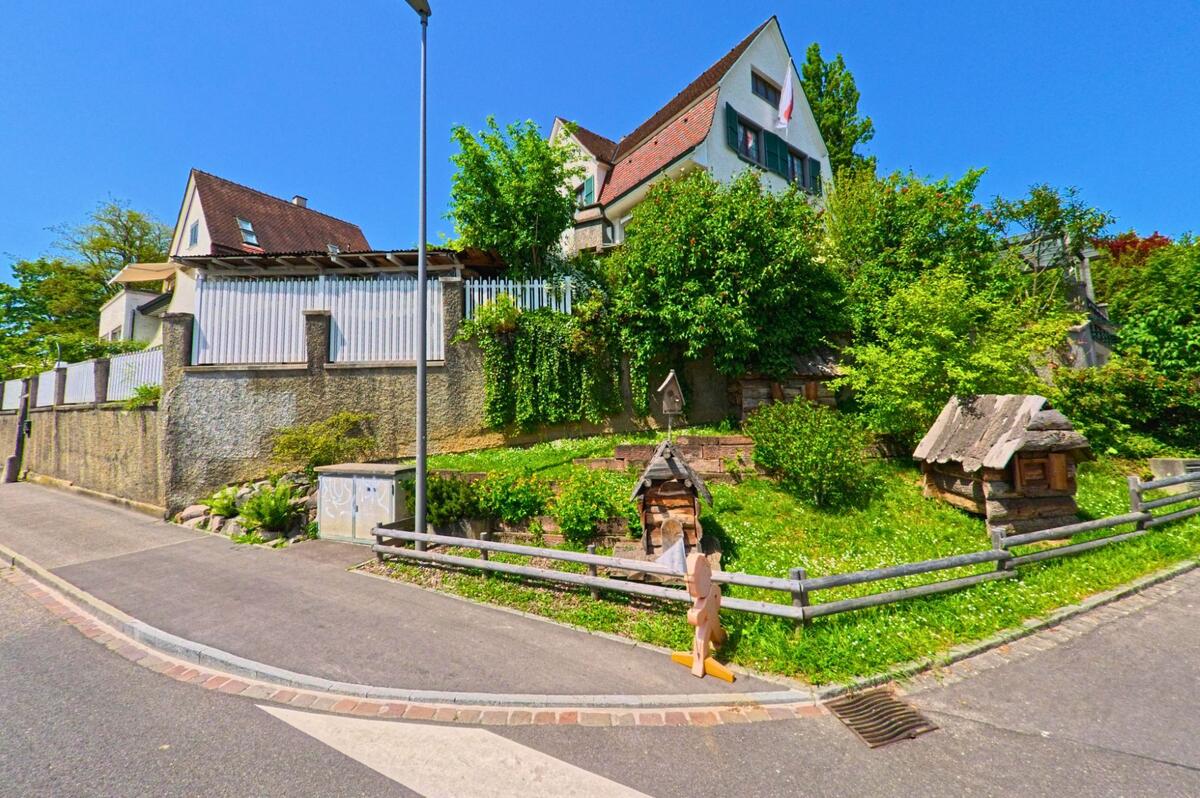 In Allschwil (4123, Kanton Baselland) liegt ein Wohnhaus mit Schweizer Fahne oberhalb eines bepflanzten Hangs, an dem kleine Holzspielhäuschen aufgestellt sind.