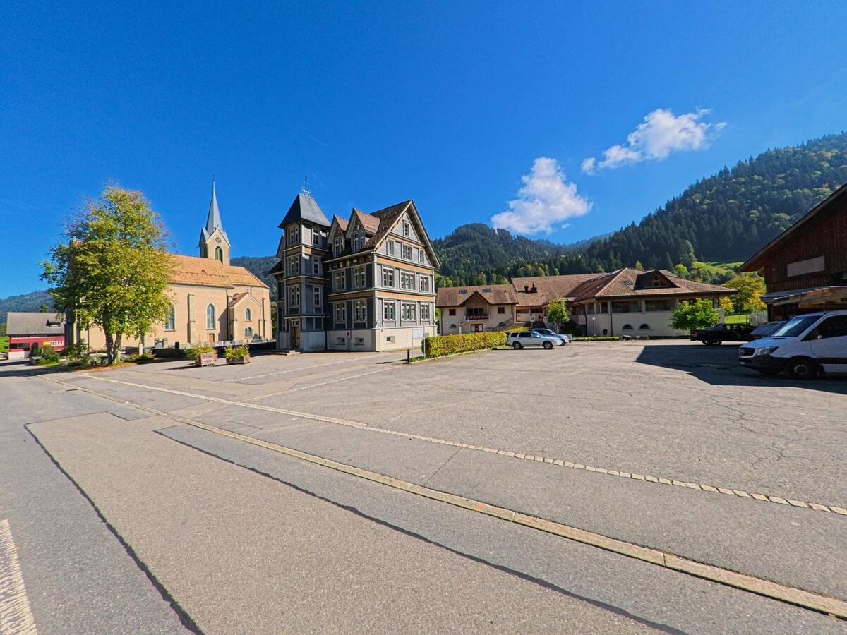 Auf dem offenen Dorfplatz stehen Kirche und Wohnhäuser vor alpiner Kulisse in Alpthal (8849, Kanton Schwyz).
