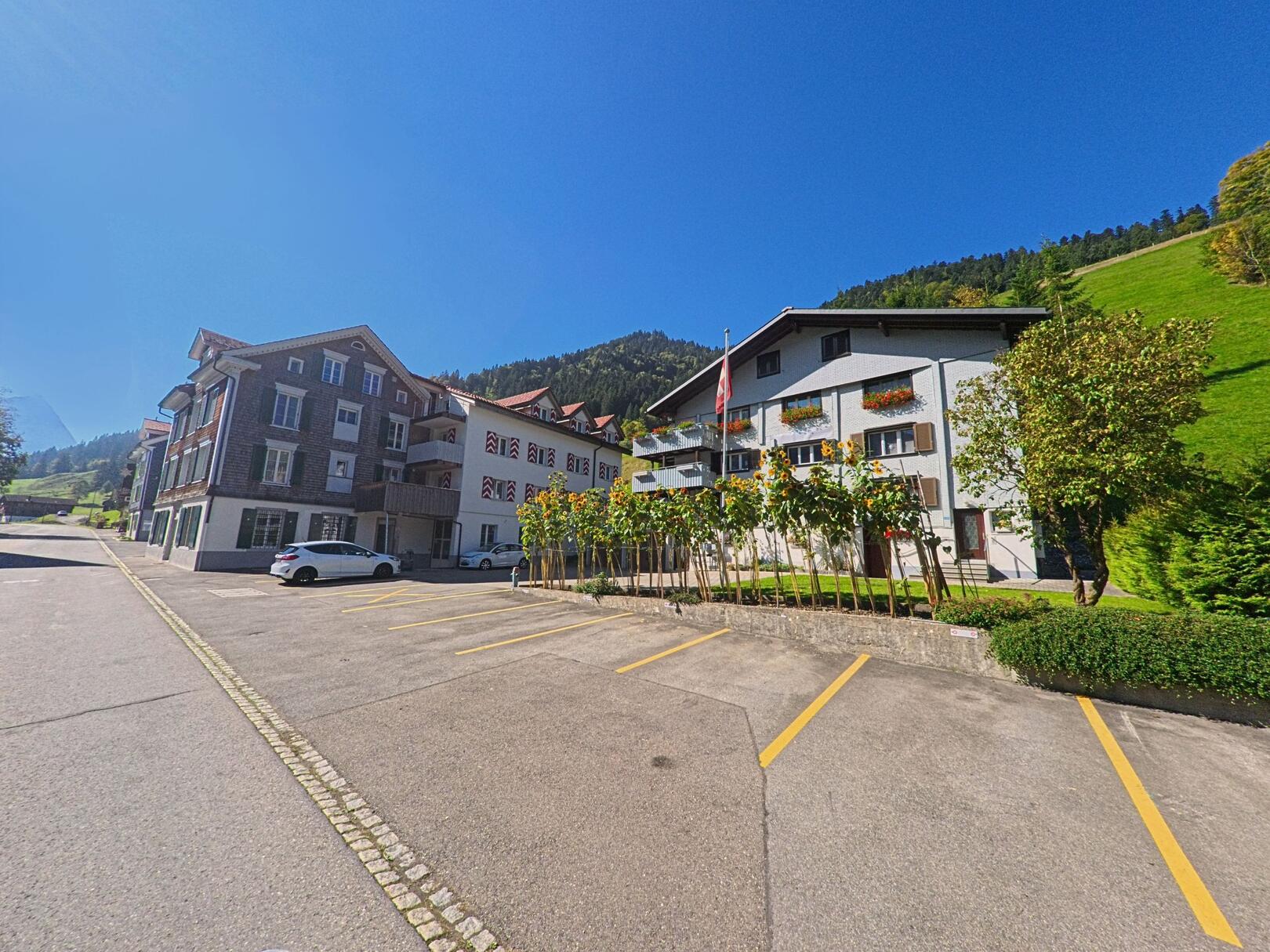 Vor den Häusern in Alphtal (8849, Kanton Schwyz) liegen unter blauem Himmel mehrere Parkplätze, während am Hang dahinter begrünte Wälder ansteigen.