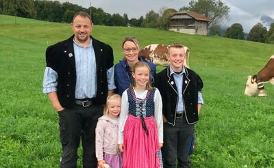 Die Familie von Kaenel aus Aeschiried (3703, Kanton Bern) steht auf einer Wiese ihrer Alpkäserei, umgeben von Kühen und Berglandschaft.