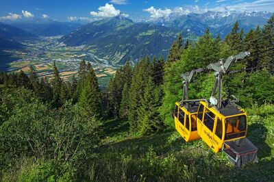 Die gelben Gondeln der Älplibahn in Malans (7208, Kanton Graubünden) fahren durch einen Wald mit Blick auf das weite Tal und die umliegenden Berge.