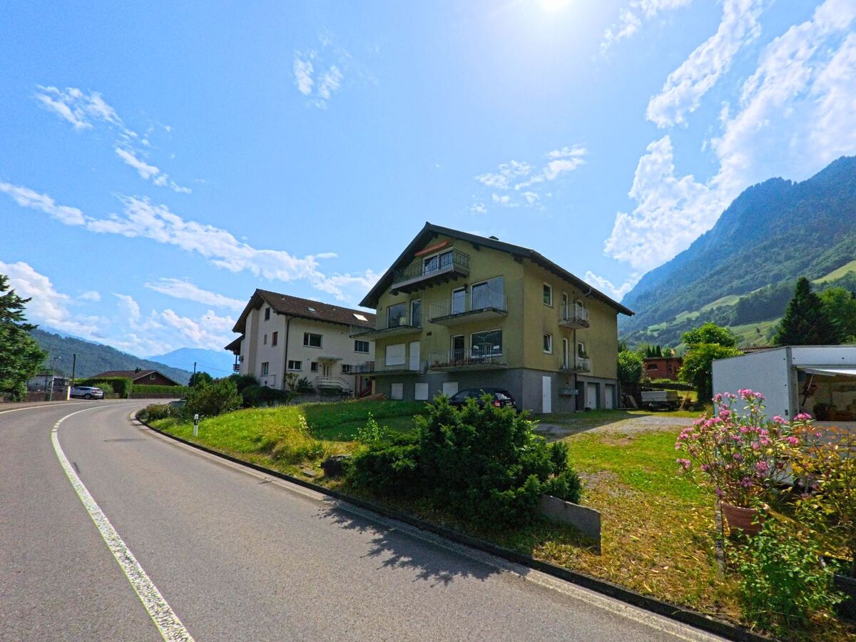 Eine sanft geschwungene Strasse fuehrt in Alpnach (6053, Kanton Obwalden) an Wohnhaeusern vorbei, waehrend im Hintergrund die bewaldeten Haenge der umliegenden Berge sichtbar sind.