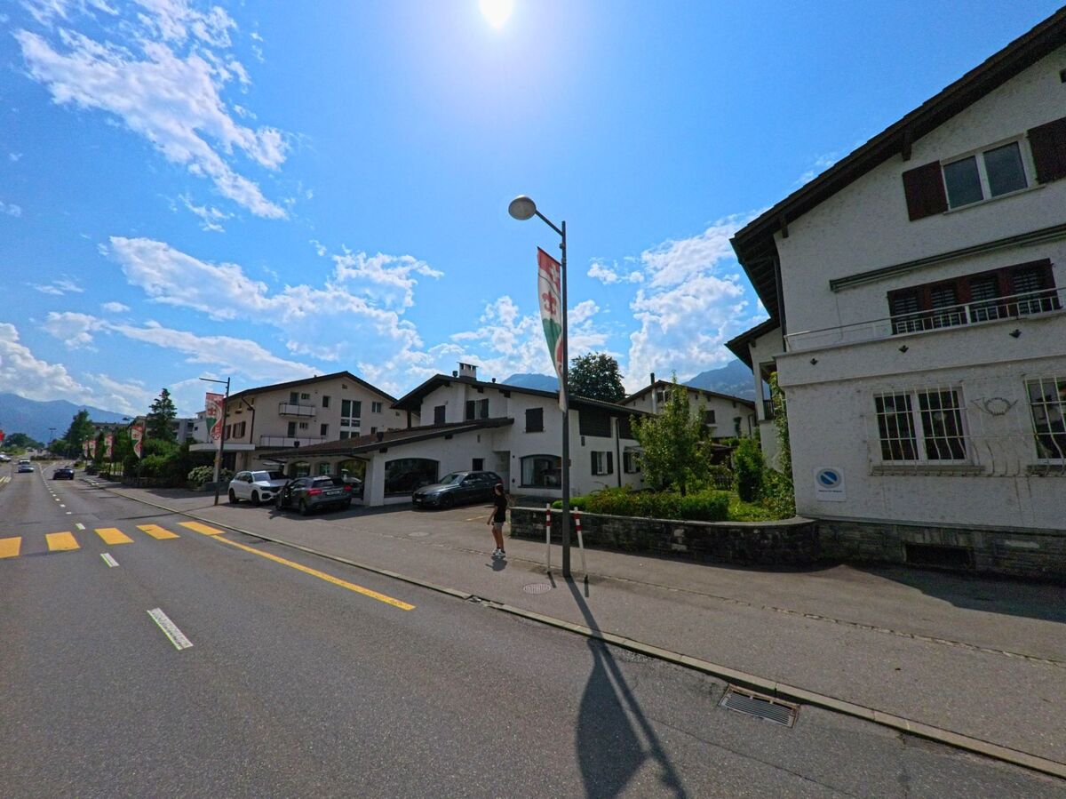 Entlang einer breiten Strasse in Alpnach (6055, Kanton Obwalden) stehen weisse Wohnhaeuser, waehrend eine Fahne mit Gemeindewappen im Sonnenlicht flattert.