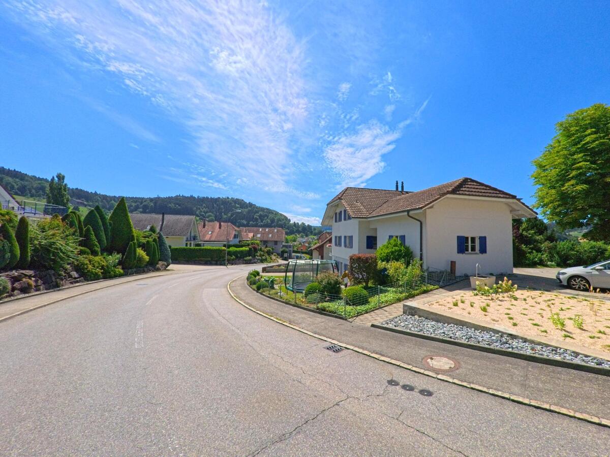 In Altbueron (PLZ 6147, Kanton Luzern) windet sich eine ruhige Wohnstrasse an gepflegten Haeusern und Gaerten vorbei, darunter ein Haus mit blauen Fensterlaeden und Kiesbeet im Vorgarten.