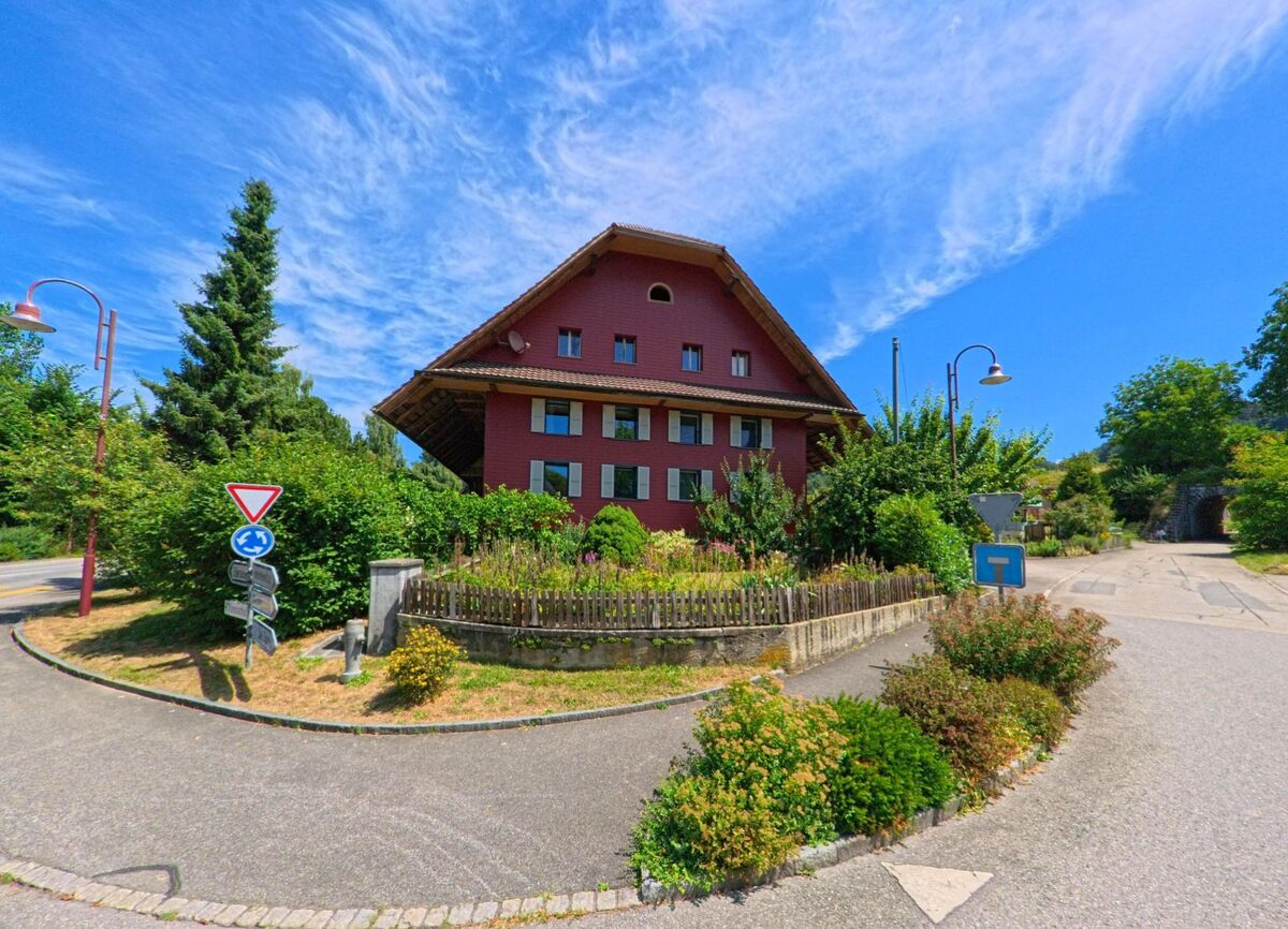 Ein grosses Haus mit roter Holzfassade steht in Altbueron (PLZ 6147, Kanton Luzern) an einer Verkehrsinsel mit Verkehrsschildern und gepflegter Bepflanzung.