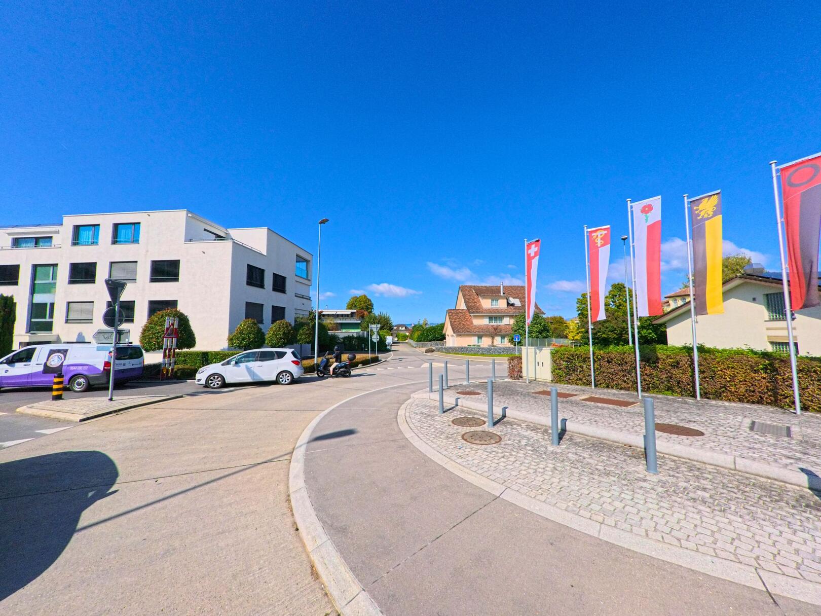 In Altendorf (8852, Kanton Schwyz) zeigt die Szene eine leicht gekruemmte Strasse mit mehreren Fahnen, modernen Gebaeuden und einem Haus im Hintergrund bei klarem Himmel.