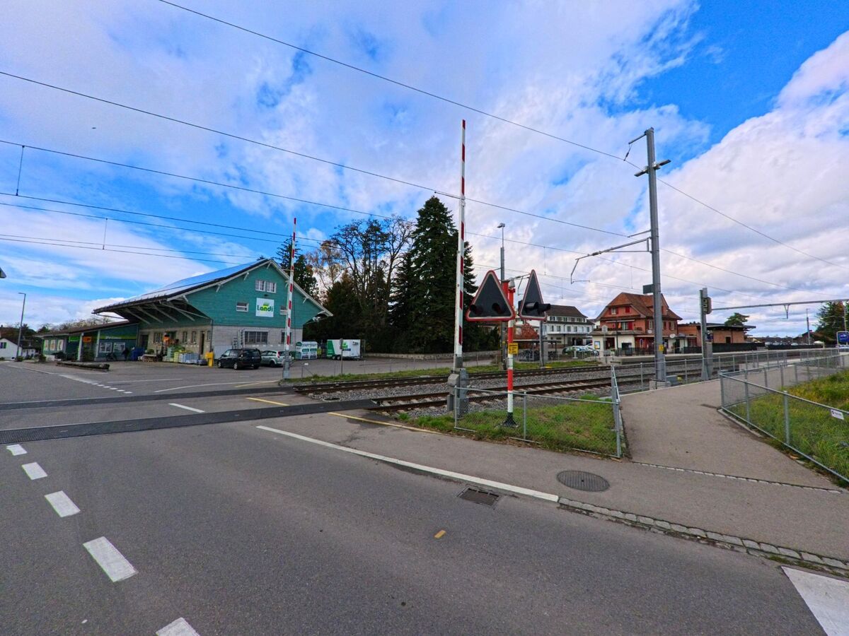Eine breite Dorfstrasse in Alterswilen (8565, Kanton Thurgau) führt zwischen Wohnhäusern und Gebäuden mit Fahnenmasten hindurch, während ein Auto am Rand parkiert.