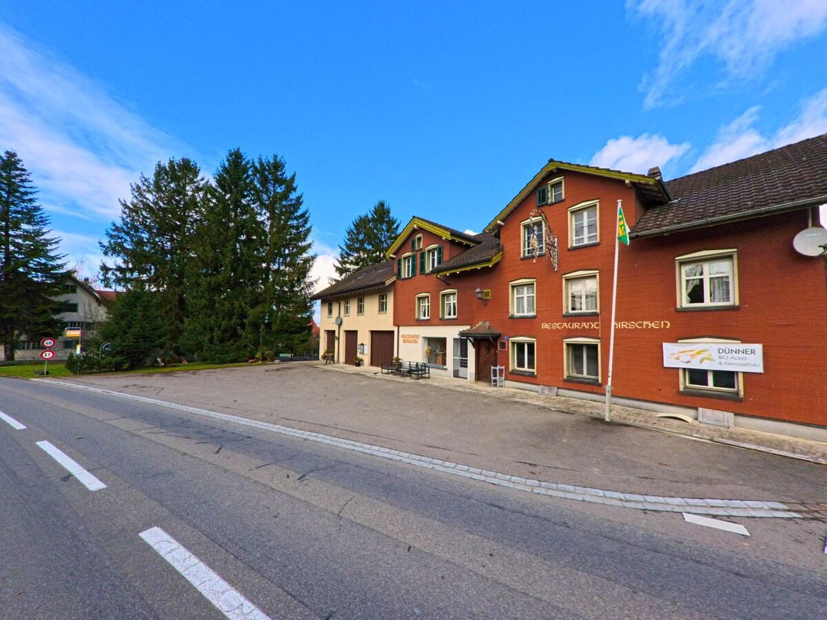 An der Hauptstrasse in Alterswilen (8565, Kanton Thurgau) steht ein rotes Gebäude mit der Aufschrift «Restaurant Hirschen» neben hohen Bäumen und einem freien Platz davor.