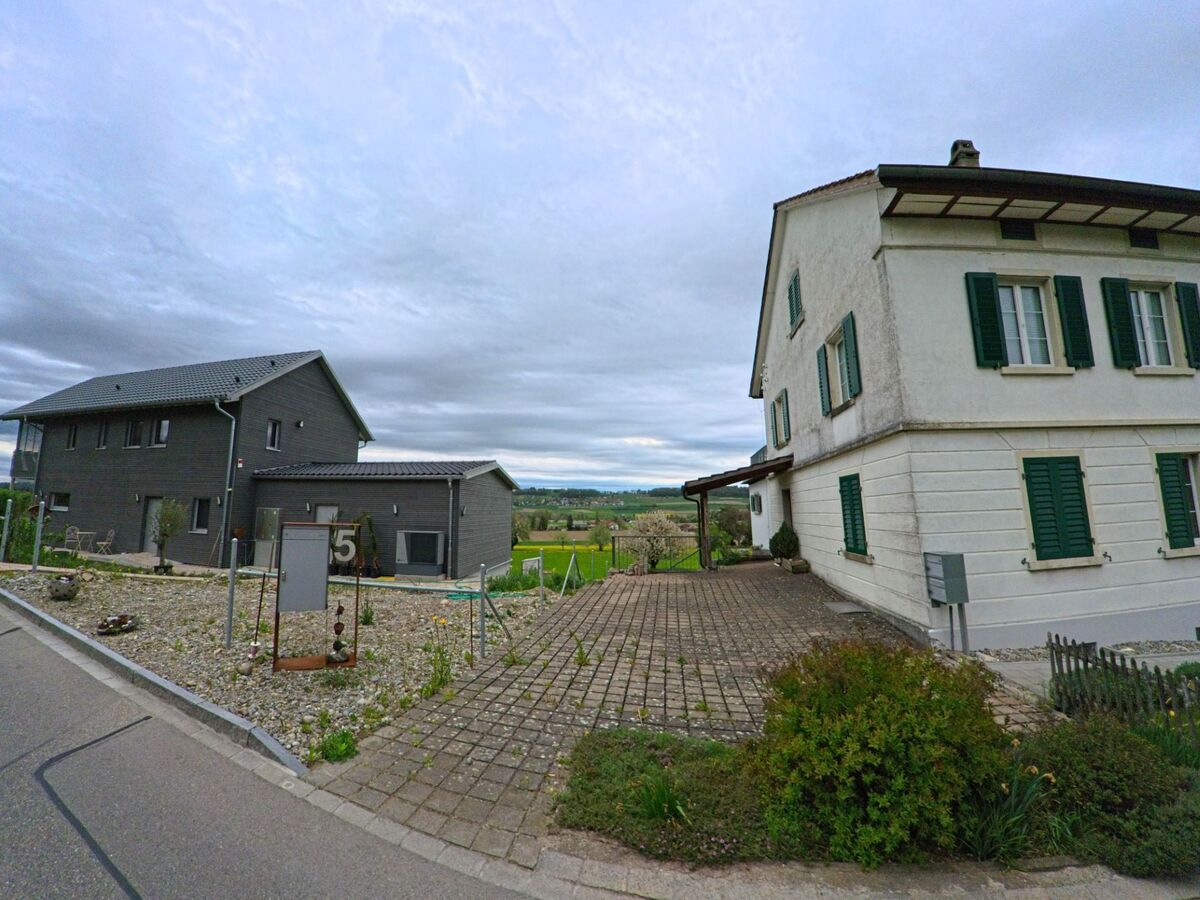 Zwei Wohnhäuser in Altikon (8479, Kanton Zürich) stehen sich gegenüber, getrennt durch einen gepflasterten Vorplatz, von dem aus der Blick in die offene Landschaft fällt.