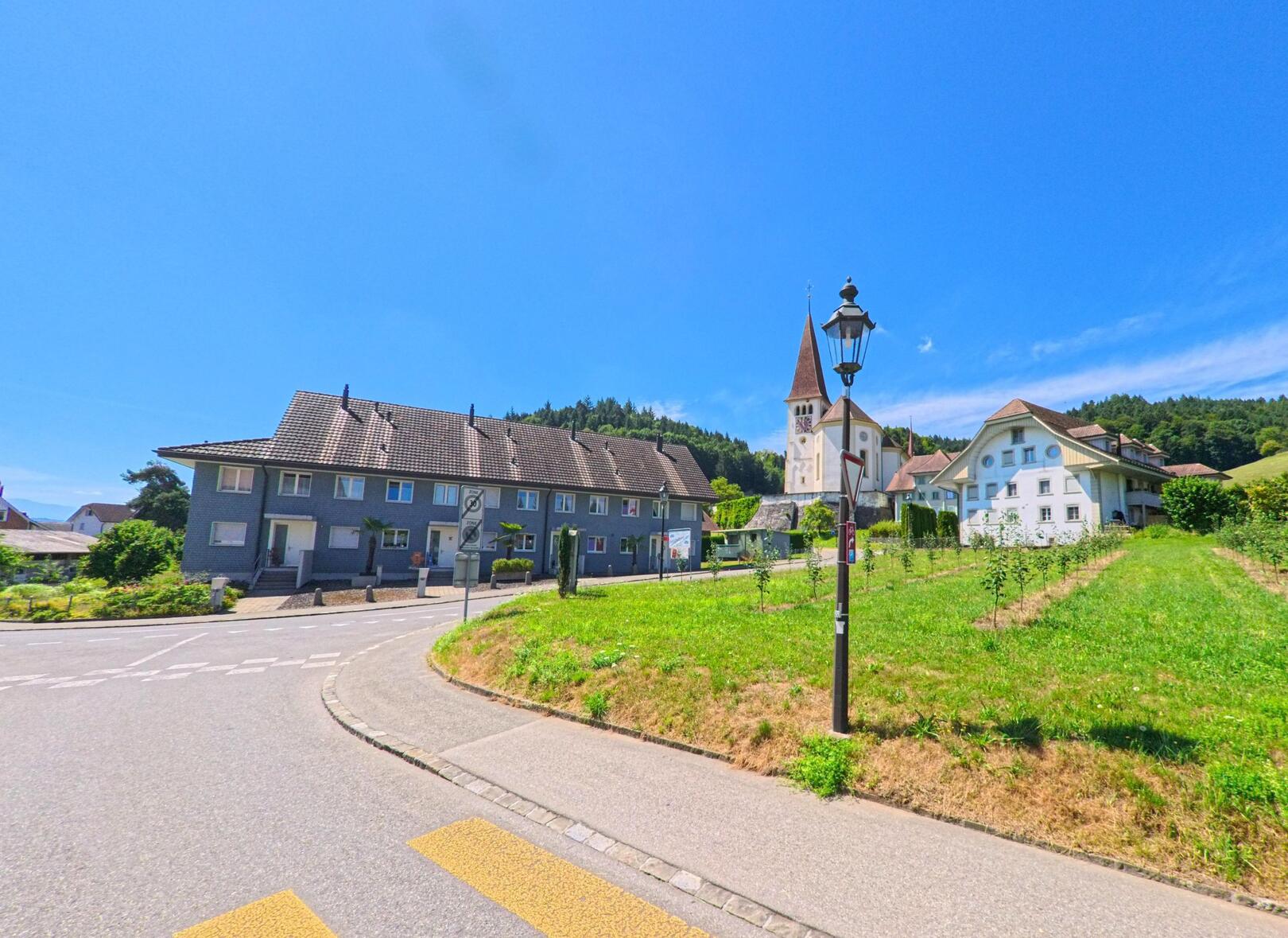 In Altishofen (PLZ 6246, Kanton Luzern) führt eine Kurve vorbei an einem Zebrastreifen, während im Hintergrund die Kirche und angrenzende Wohnhäuser von Obstgärten und Wiesen umgeben sind.