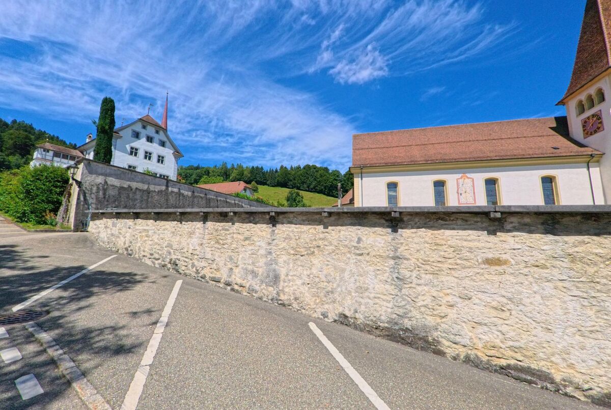 In Altishofen (PLZ 6246, Kanton Luzern) grenzen ein Kirchengebaeude mit Uhrturm und ein weiteres historisches Haus an eine hohe Steinmauer bei einem leeren Parkplatz.