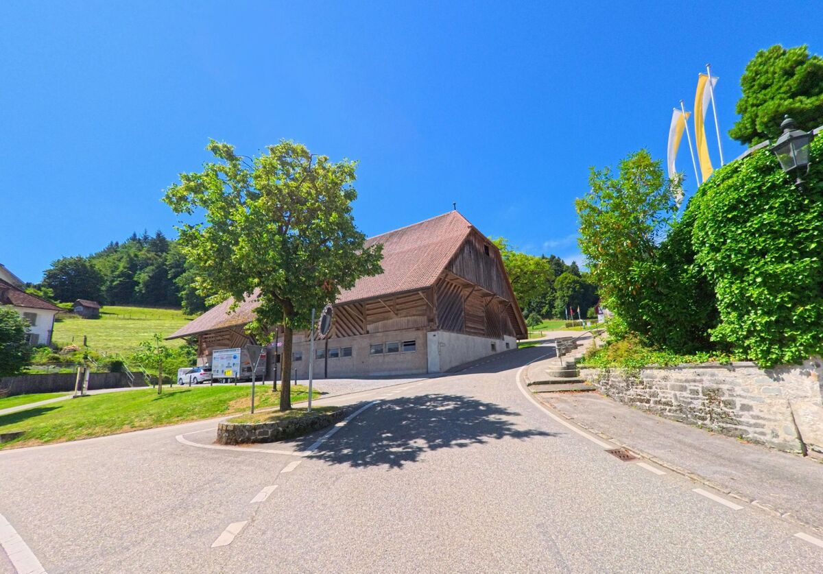 Ein breiter Weg mit Kurve fuehrt in Altishofen (PLZ 6246, Kanton Luzern) an einer grossen Scheune vorbei, flankiert von Wiesen, Baeumen und einer mit Efeu bewachsenen Mauer.