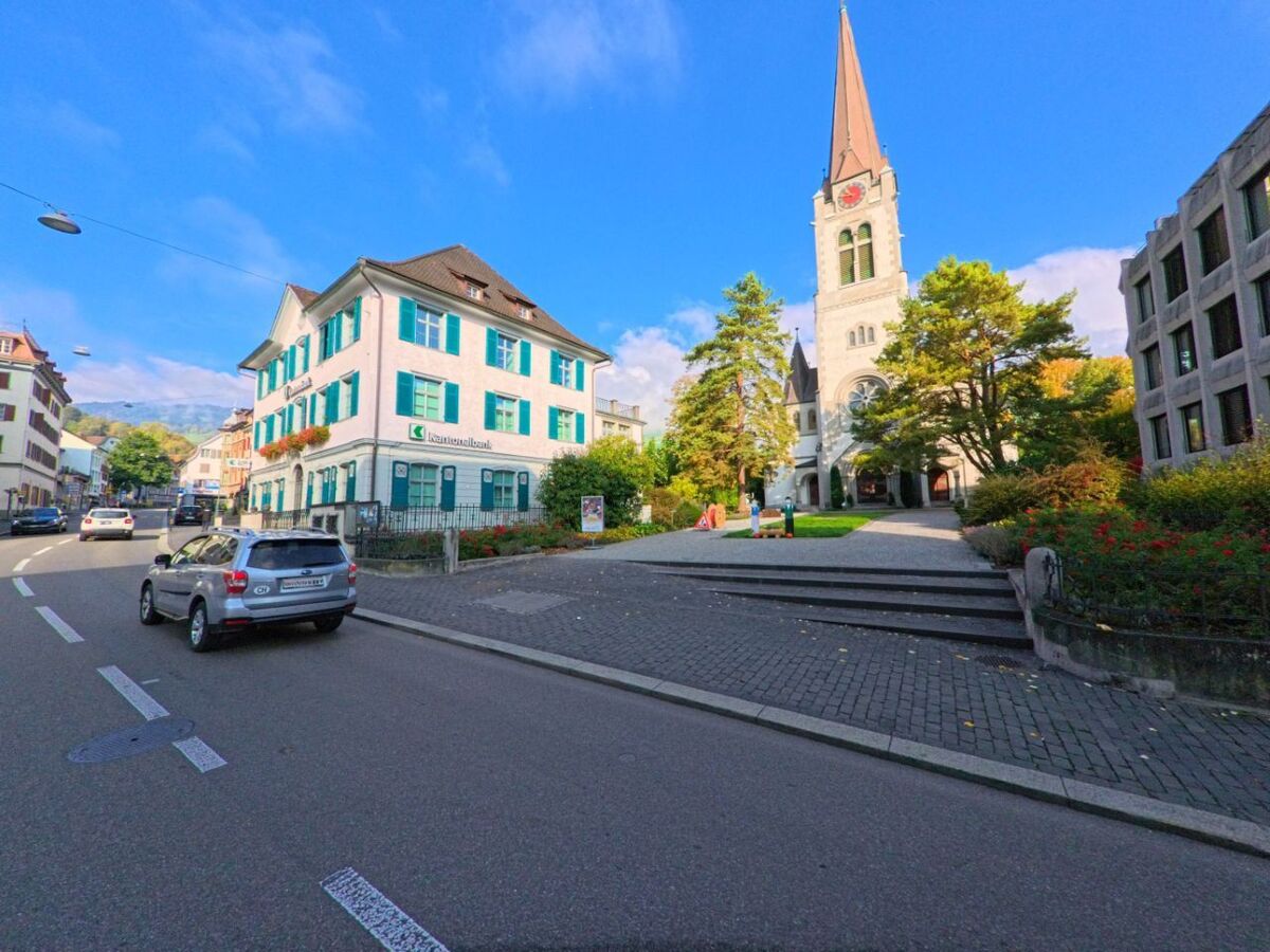 Neben einem hellen Gebäude mit grünen Fensterläden steht in Altstätten (9450, Kanton St. Gallen) eine Kirche mit hohem Turm an einer befahrenen Strasse unter blauem Himmel.