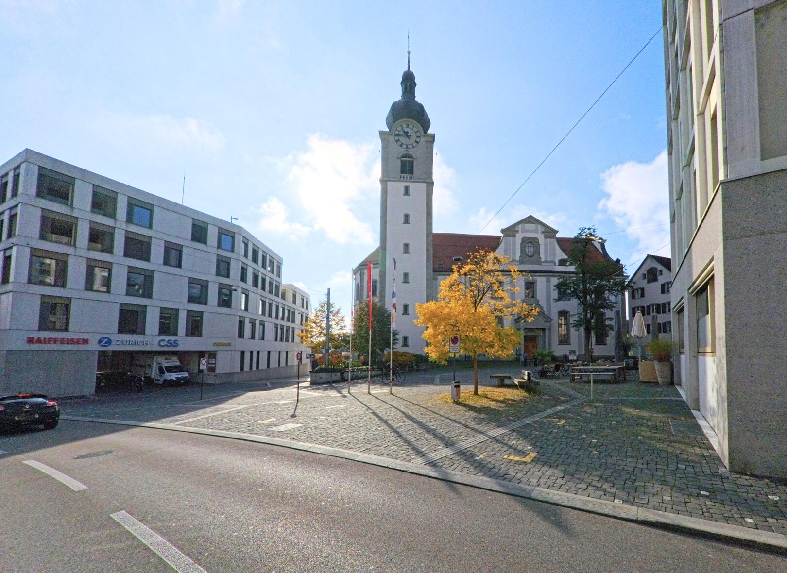 Auf einem gepflasterten Platz in Altstätten (9450, Kanton St. Gallen) ragt ein Kirchturm mit Uhr vor blauem Himmel empor, daneben leuchtet ein Baum im Herbstlicht.