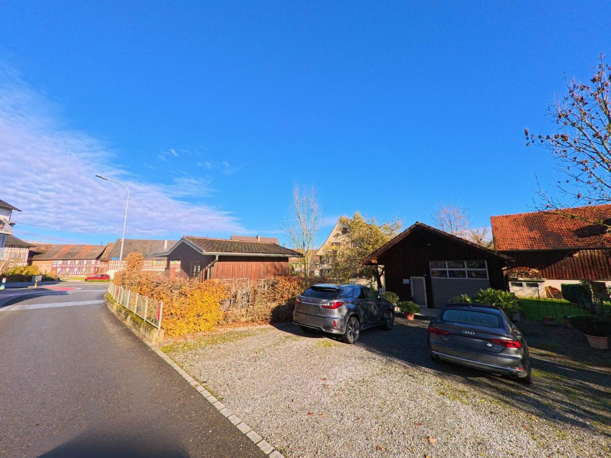 Auf einem Kiesparkplatz neben Holzgebaeuden und Wohnhaeusern zeigt sich eine ruhige Wohnlage in Amlikon-Bissegg (8514, Kanton Thurgau) bei klarem Herbsthimmel.