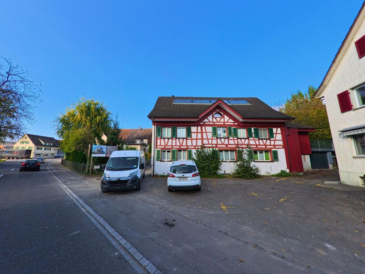 Vor einem markanten Fachwerkhaus mit roten Balken stehen Fahrzeuge auf einem kleinen Parkplatz in Amriswil (8580, Kanton Thurgau) unter klarem Himmel.