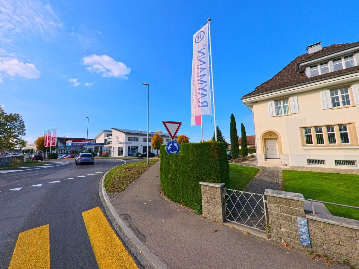 An einem Kreisverkehr in Amriswil (8580, Kanton Thurgau) stehen Wegweiser, Fahnenmasten und Wohnhaeuser entlang einer Strasse mit gelbem Zebrastreifen.