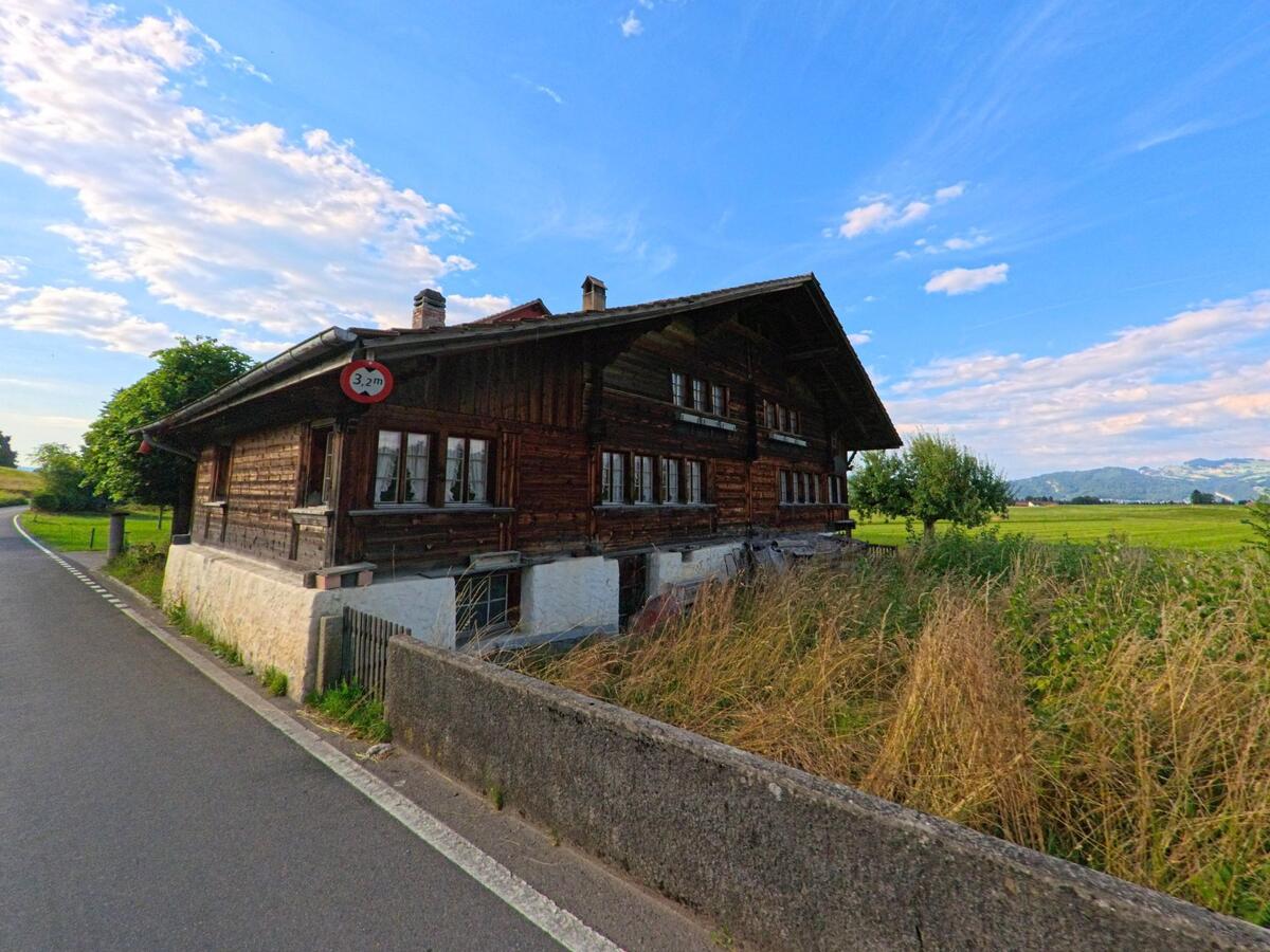 Ein traditionelles Bauernhaus aus Holz steht in Amsoldingen (3633, Kanton Bern) am Rand einer schmalen Landstrasse, umgeben von Wiesen und freiem Blick in die Hügellandschaft.