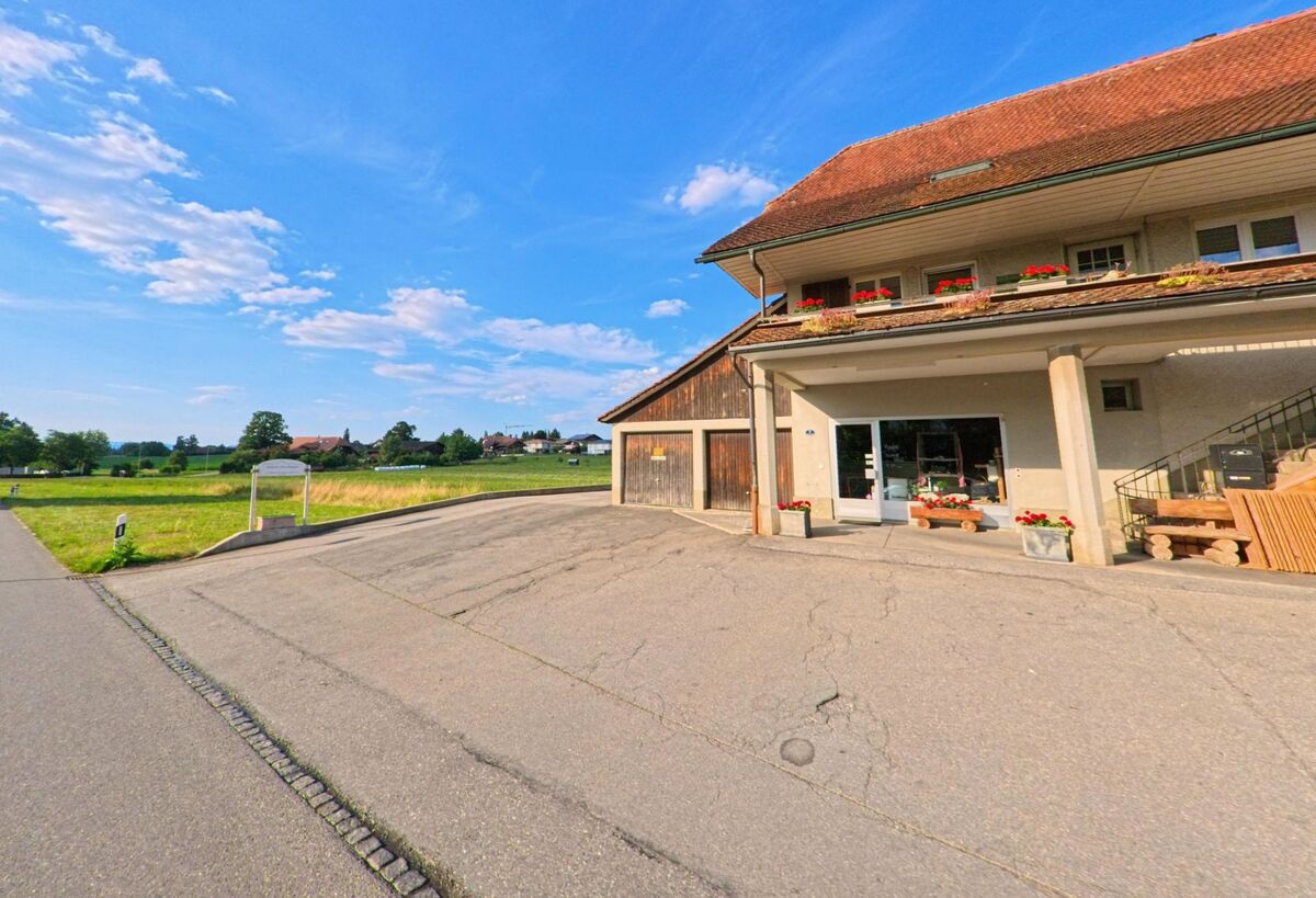 In Amsoldingen (3633, Kanton Bern) befindet sich ein Hofladen mit überdachtem Eingang in einem Bauernhaus, das an eine leicht ansteigende Landstrasse grenzt und von offenen Feldern umgeben ist.