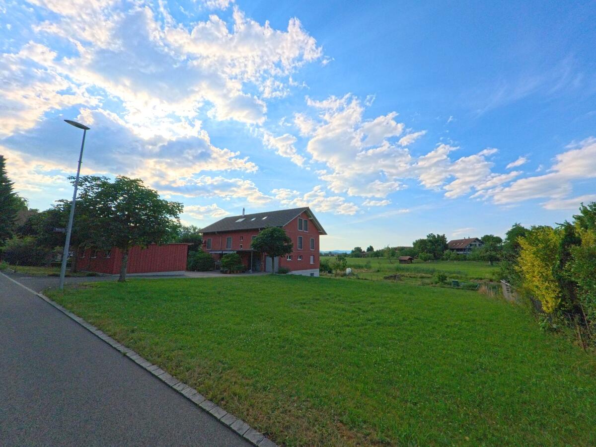 Am Rande von Amsoldingen (3633, Kanton Bern) steht ein rotes Wohnhaus neben einer grossen Wiese, mit Blick auf verstreut liegende Gebäude und Gartenanlagen in ländlicher Umgebung.