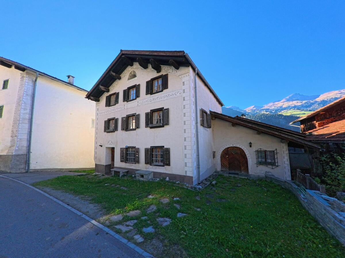 Am Rand einer schmalen Strasse in Andeer (7440, Kanton Graubünden) steht ein historisches Wohnhaus mit dekorativer Fassade und Holzläden vor alpiner Bergkulisse.