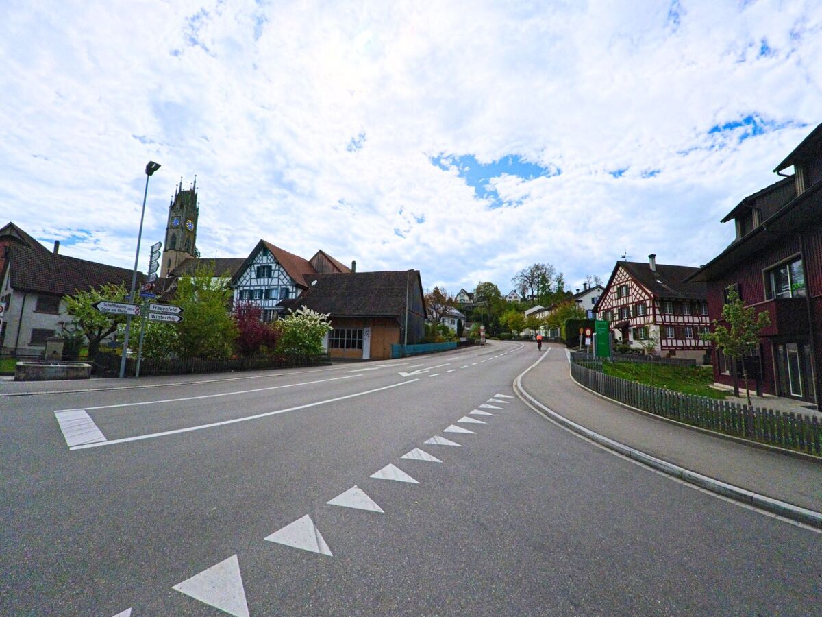  Im Zentrum von Andelfingen (8453, Kanton Zürich) verläuft eine leicht ansteigende Strasse zwischen Fachwerkhäusern, mit dem markanten Kirchturm im Hintergrund.