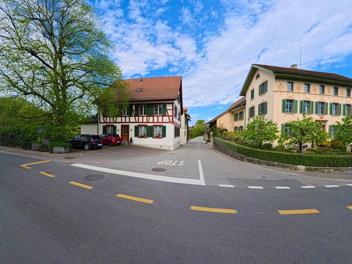  In Andelfingen im Kanton Zürich (PLZ 8453) stehen an einer ruhigen Strassengabelung ein traditionelles Fachwerkhaus und ein historisches Gebäude mit grünen Fensterläden.