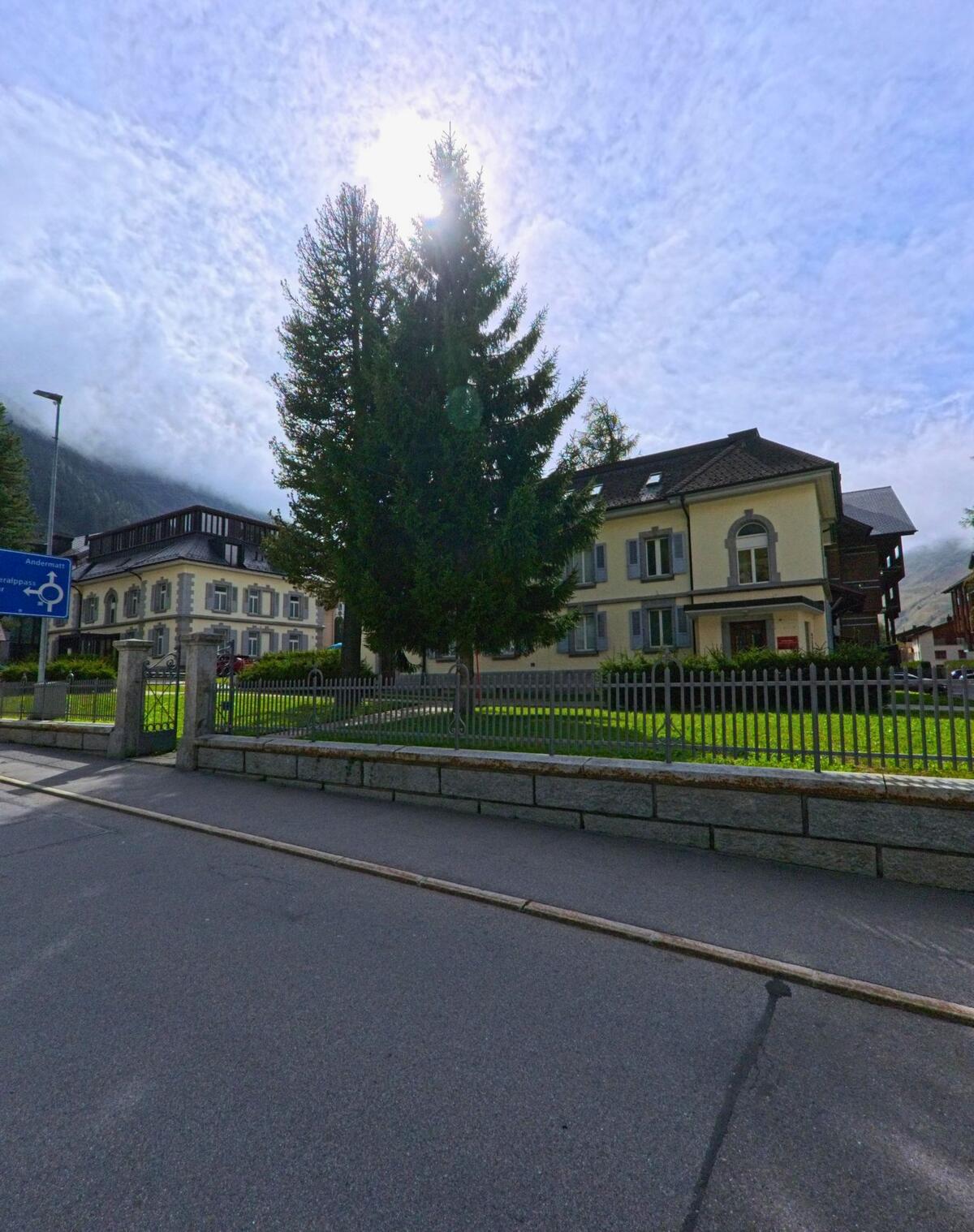Vor historischen Wohnhaeusern in Andermatt (6490, Kanton Uri) steht eine grosse Tanne, waehrend die Sonne hinter Wolken hervorscheint.