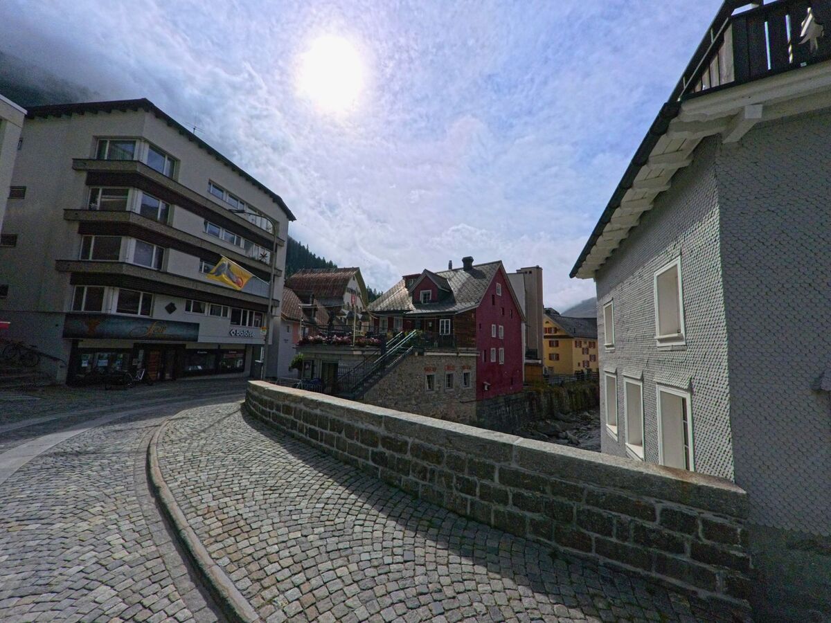 Eine gepflasterte Strasse in Andermatt (6490, Kanton Uri) fuehrt an modernen und traditionellen Gebaeuden entlang, waehrend die Sonne durch die Wolken scheint.
