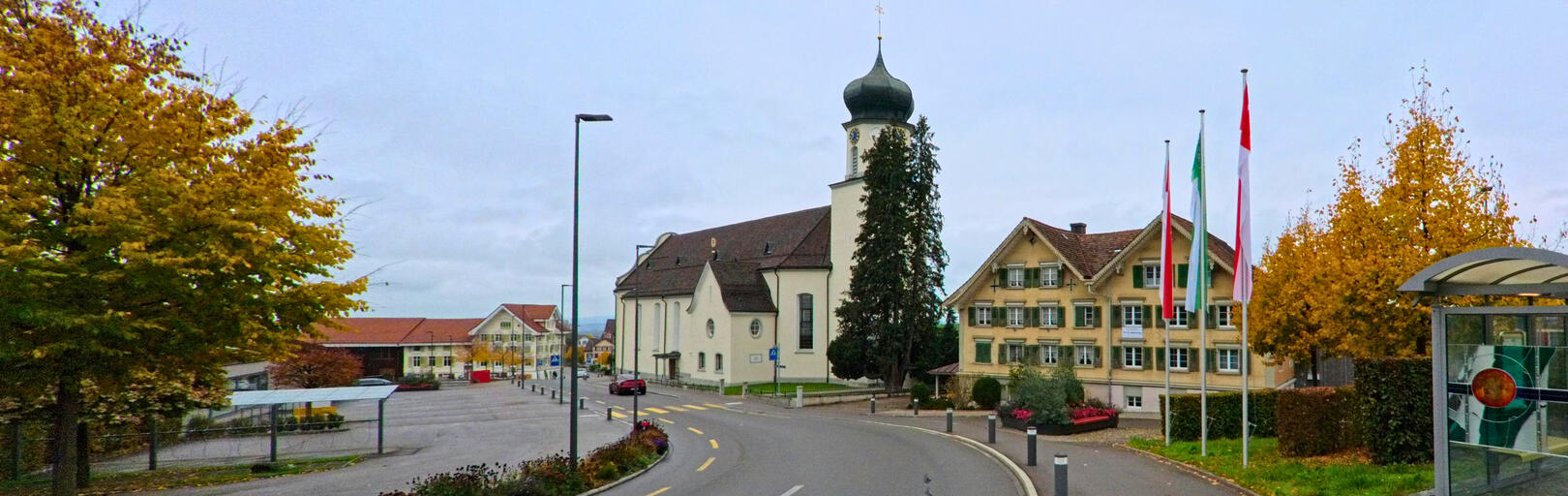 Der Blick auf die Hauptstrasse in Andwil (9212, Kanton St. Gallen) zeigt die markante Kirche und das historische Gebäude, umgeben von herbstlich gefärbten Bäumen.