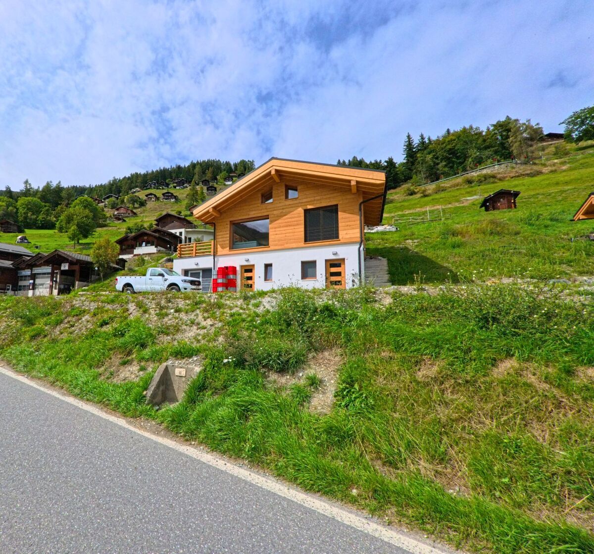 Ein modernes Chalet aus Holz steht am Hang in Anniviers (3961, Kanton Wallis), umgeben von grünen Wiesen und weiteren verstreut liegenden Hausern.