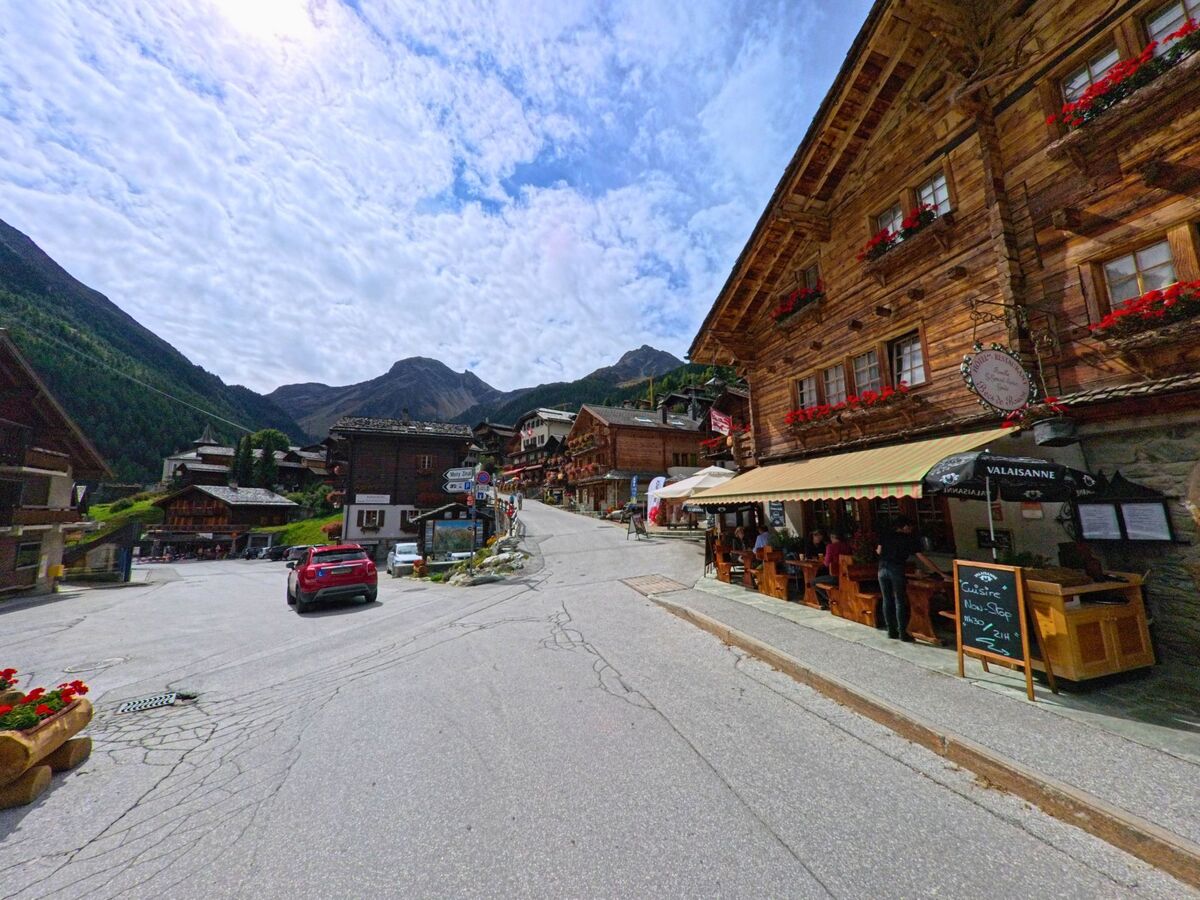 In der Dorfstrasse von Anniviers (3961, Kanton Wallis) sitzen Gaeste vor einem traditionellen Holzhaus mit Restaurant, waehrend im Hintergrund die Berggipfel sichtbar sind.
