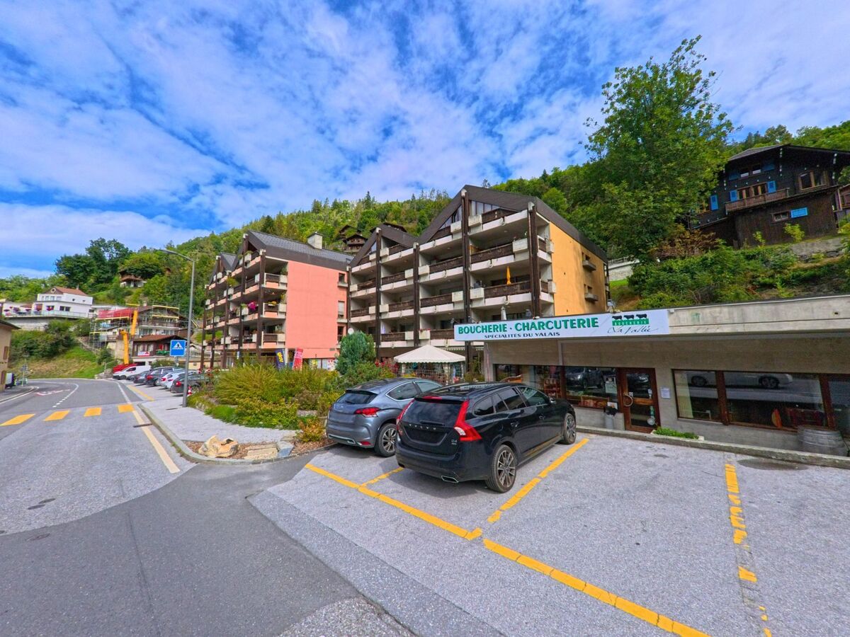 In Anniviers (3961, Kanton Wallis) stehen moderne Wohnhaeuser mit Balkonen neben einem Geschäft für Fleischwaren, im Vordergrund sind Parkplätze und die Strasse sichtbar.