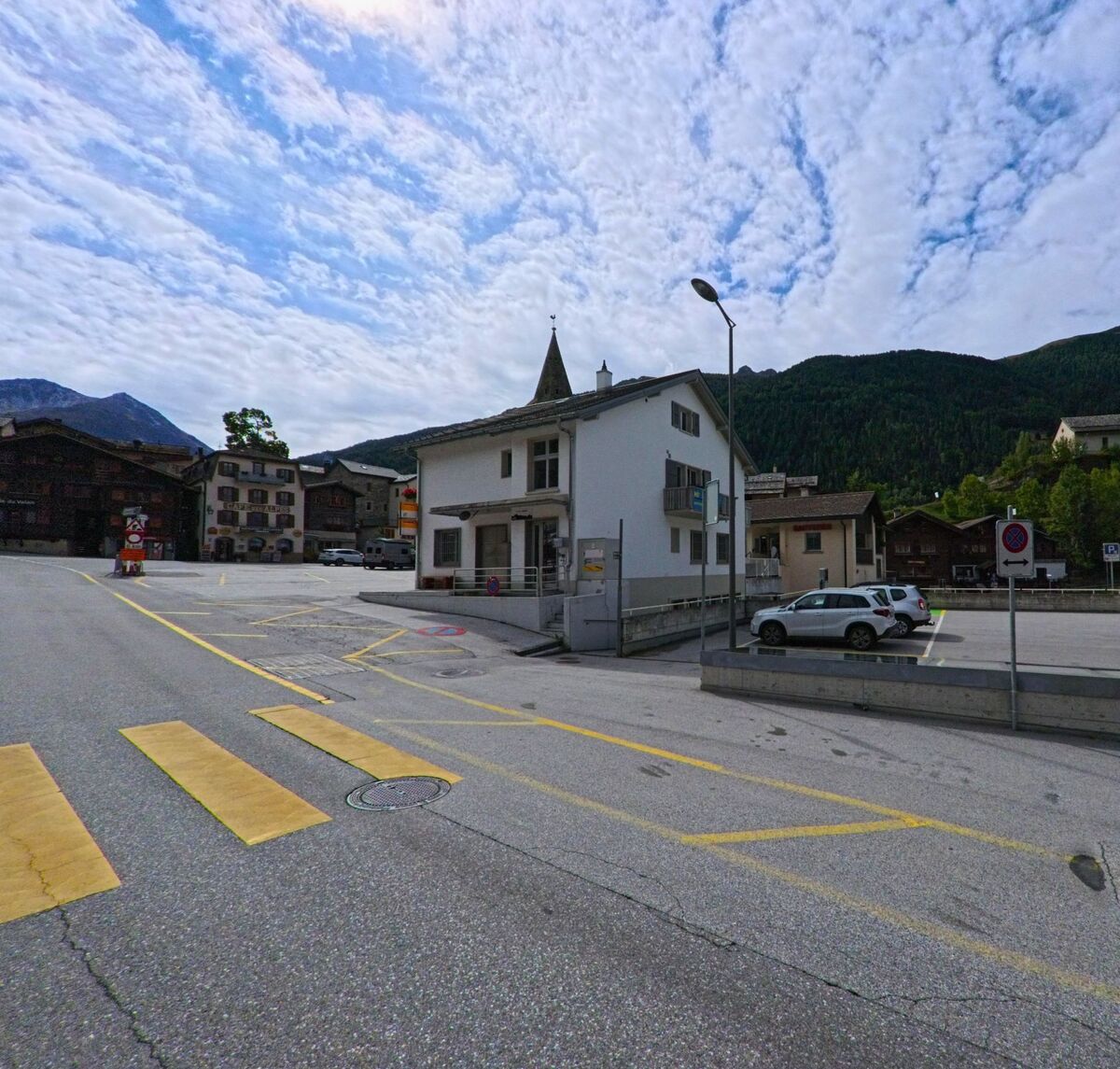 Ein kleines weisses Gebaeude mit Parkplatz liegt im Zentrum von Anniviers (3961, Kanton Wallis), im Hintergrund ragt der Kirchturm vor den bewaldeten Berghaengen auf.