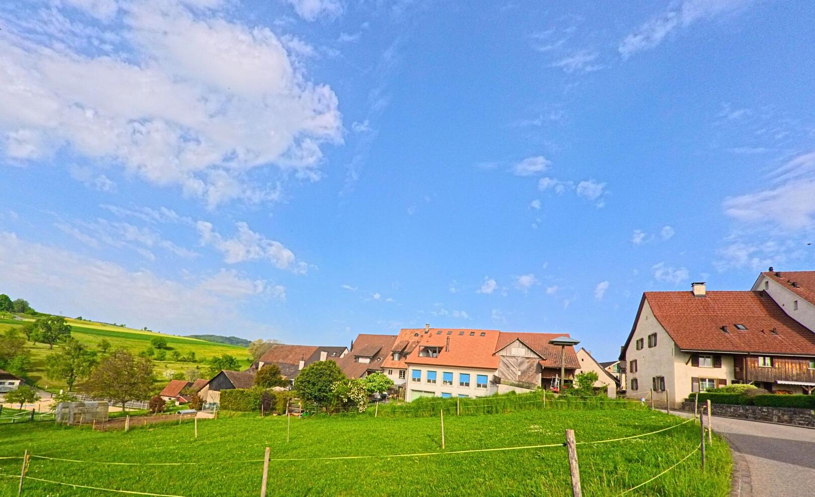 Am Rand von Anwil (4469, Kanton Baselland) liegt ein ruhiges Wohnquartier mit traditionellen Häusern und grünen Wiesen unter leicht bewölktem Himmel.