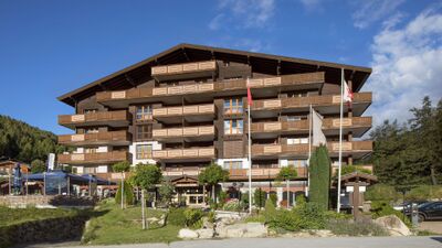 Das Aparthotel Ambassador in Bellwald (3997, Kanton Wallis) präsentiert sich mit einer grossen Holzfassade, mehreren Balkonen und einer sonnigen Terrasse vor alpiner Bergkulisse.