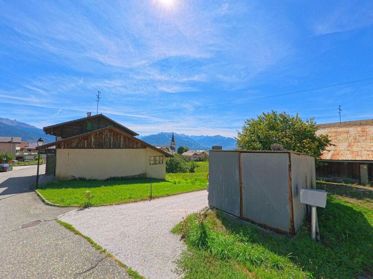 In Arbaz (1974, Kanton Wallis) stehen einfache Dorfhaeuser und Scheunen, dahinter ragt ein Kirchturm vor den umliegenden Alpen empor.
