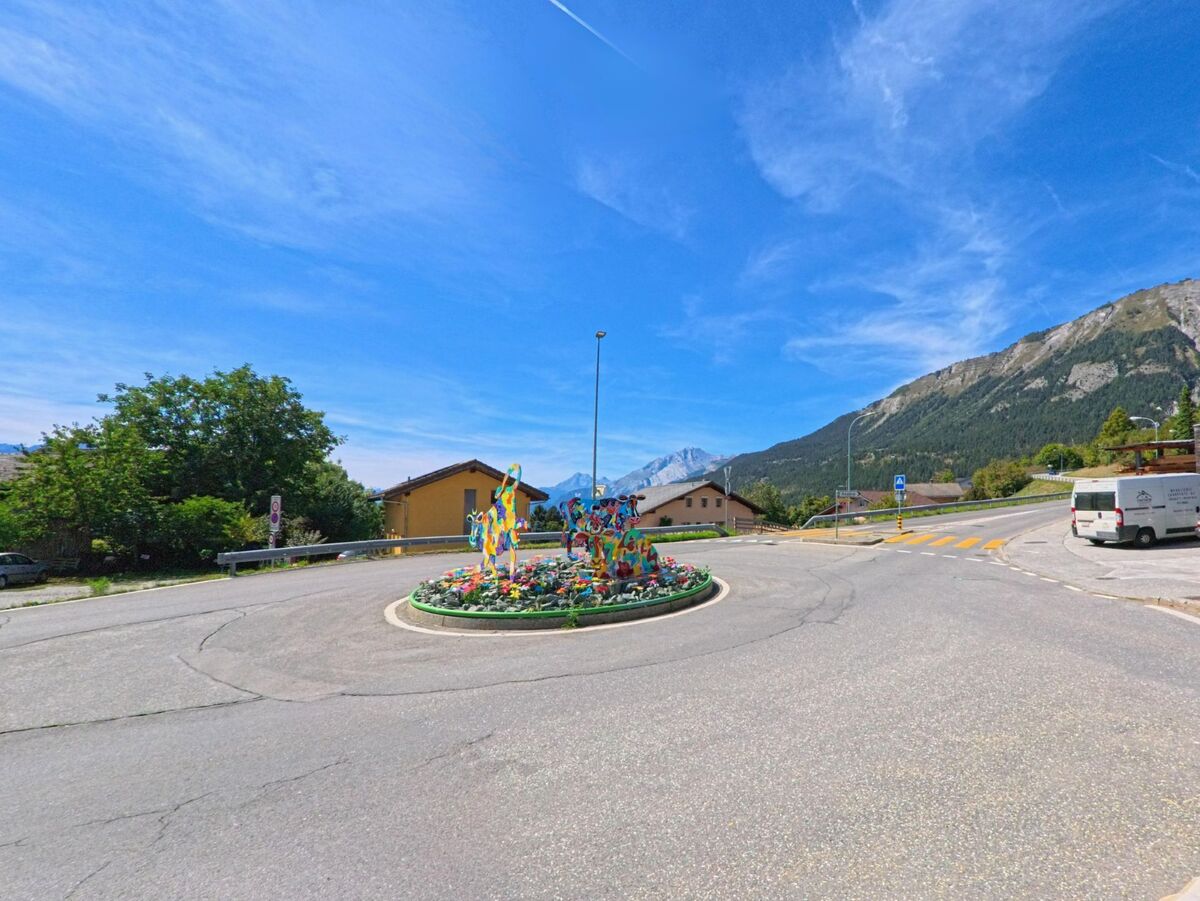 Ein Kreisverkehr in Arbaz (1974, Kanton Wallis) ist mit farbenfrohen Skulpturen geschmueckt, im Hintergrund erheben sich steile Berghaenge.