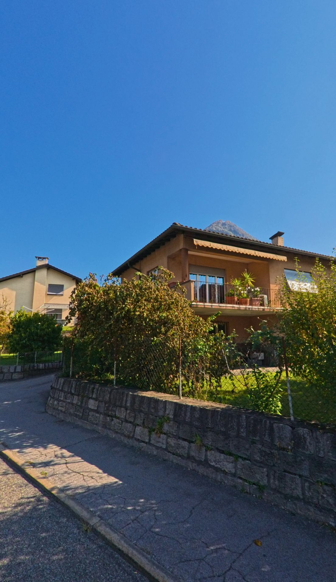 An einer ruhigen Wohnstrasse in Arbedo-Castione (6532, Kanton Tessin) steht ein Einfamilienhaus mit Balkon und Garten hinter einer niedrigen Steinmauer.