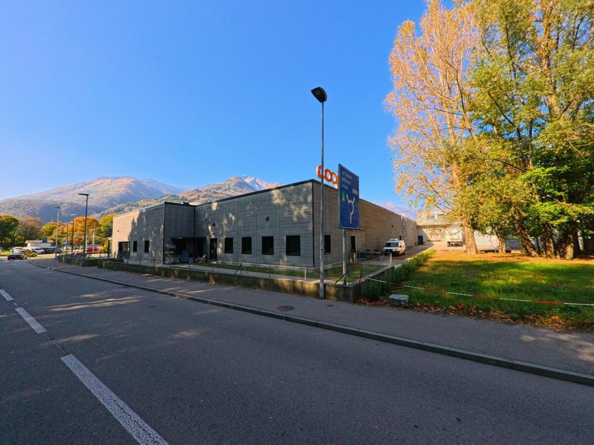 Entlang einer Strasse in Arbedo-Castione (6532, Kanton Tessin) steht ein flaches Gewerbegebäude mit angrenzender Grünfläche, im Hintergrund sind herbstliche Bäume und Berge sichtbar.