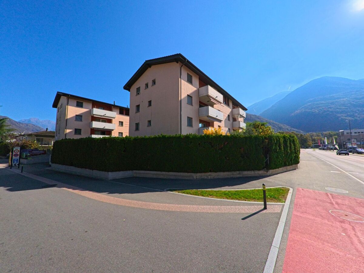An der Ecke einer ruhigen Strasse in Arbedo-Castione (6532, Kanton Tessin) stehen mehrgeschossige Wohnbauten hinter einer hohen Hecke, flankiert von Fahrbahn und Velostreifen.