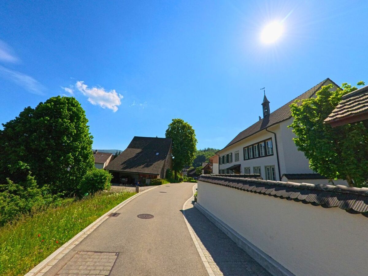 In Arboldswil (4424, Kanton Basel-Landschaft) verläuft eine schmale Strasse an einer weissen Mauer und einem Gebäude mit Türmchen unter strahlender Sonne vorbei.