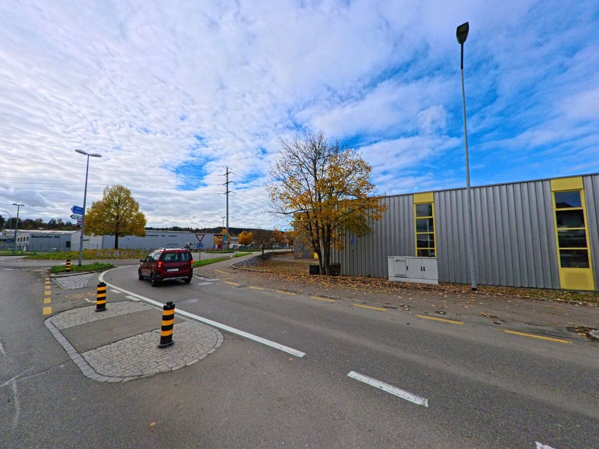 An einer verkehrsberuhigten Abzweigung in Arbon (9320, Kanton Thurgau) faehrt ein Fahrzeug vorbei an Gewerbebauten und herbstlichen Baeumen.