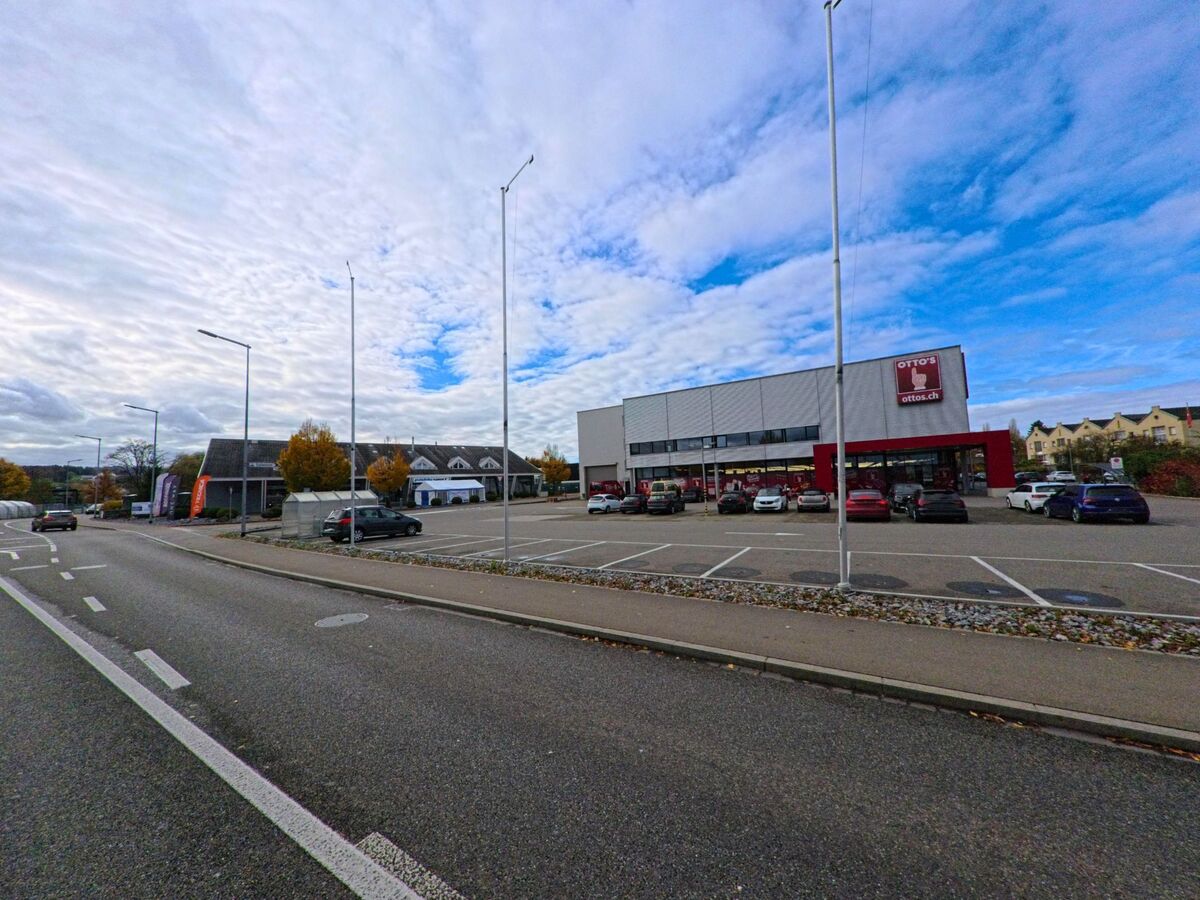 Gegenueber eines grossen Parkplatzes in Arbon (9320, Kanton Thurgau) befindet sich ein Einkaufsgebaeude entlang einer mehrspurigen Strasse unter wolkigem Himmel.