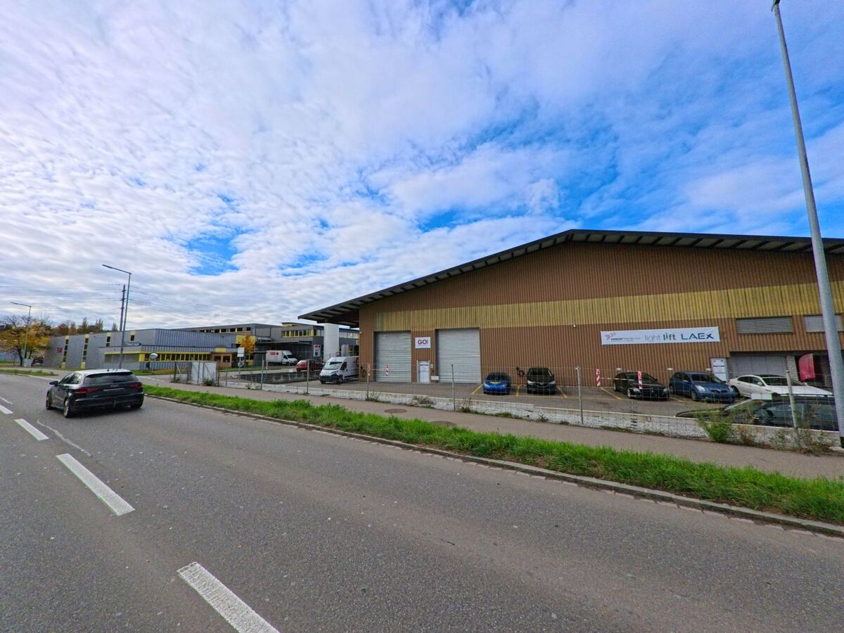 Entlang einer Hauptstrasse in Arbon (9320, Kanton Thurgau) stehen Gewerbehallen mit Parkplaetzen und abgestellten Fahrzeugen unter wechselhaftem Himmel.